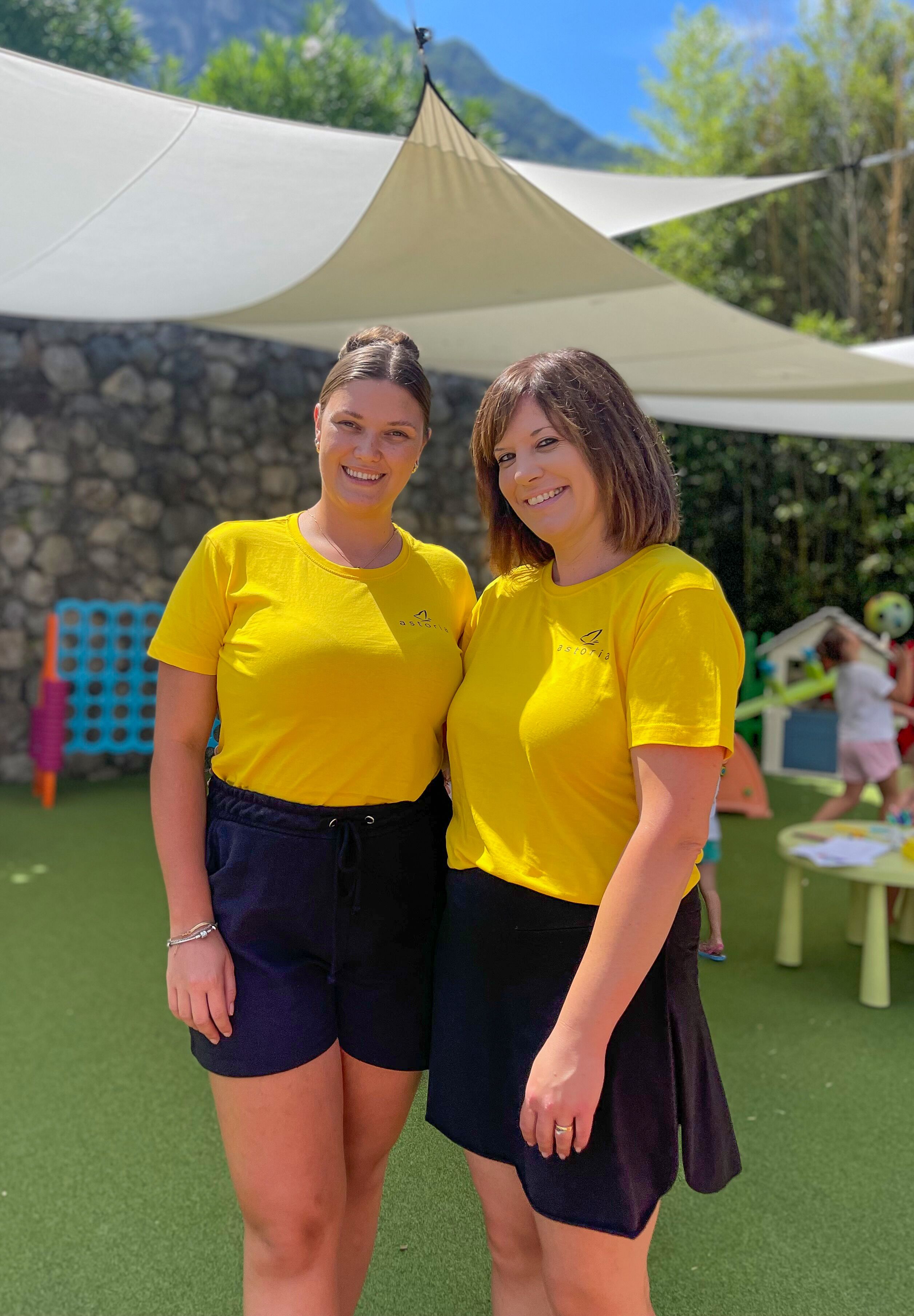 Two people in yellow shirts standing on a green surface with a stone wall and a tent in the background.