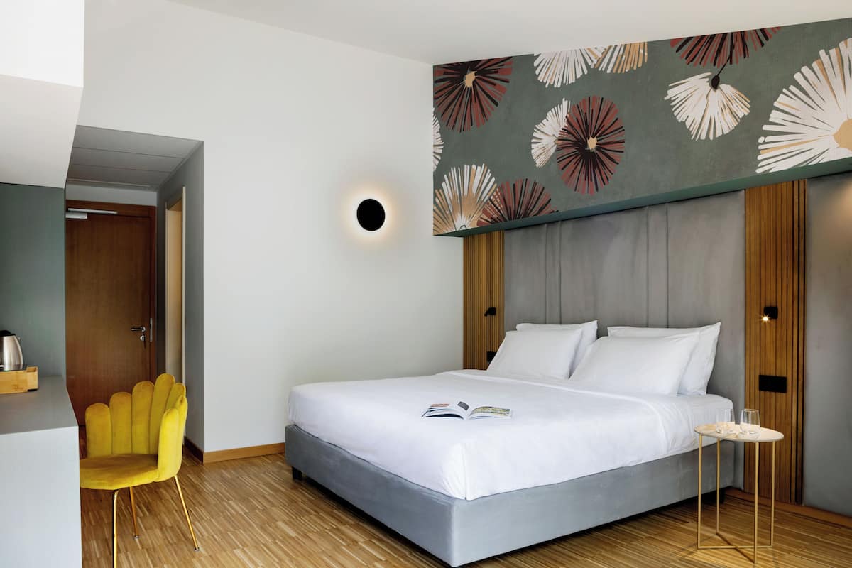 A modern hotel room with a large bed, a small side table, a yellow armchair, and a decorative wall with a floral pattern.