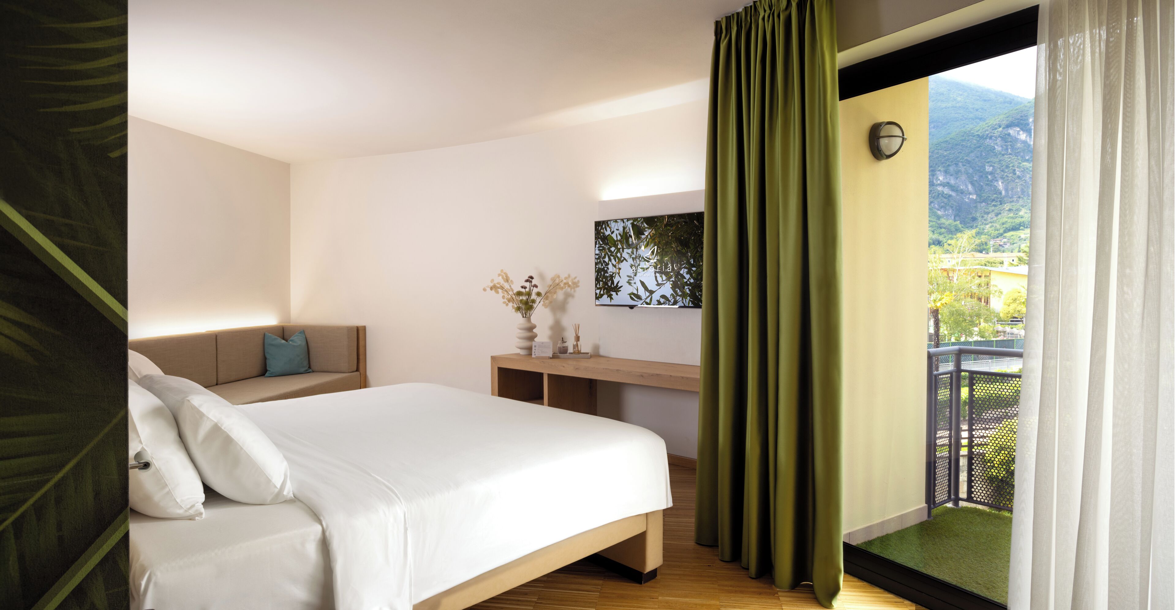 Habitación de hotel con cama, escritorio, silla, una pequeña mesa con un florero, y vistas a zonas verdes y montañas a través de la ventana.