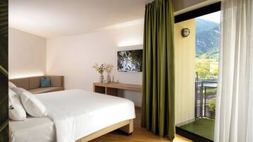 Habitación de hotel con cama, escritorio, silla, una pequeña mesa con un florero, y vistas a zonas verdes y montañas a través de la ventana.