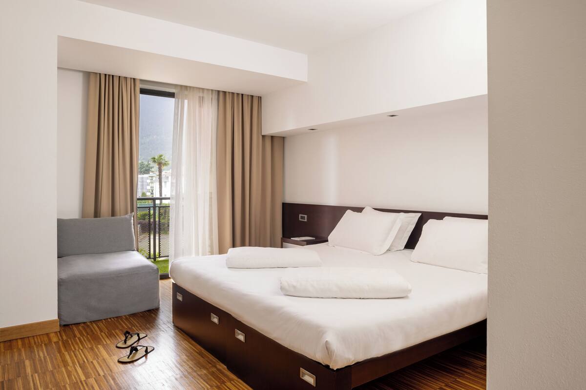 A hotel room with a bed, a grey armchair, and a view of a balcony with curtains.