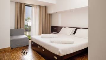 Una habitación de hotel con una cama, un sillón gris y vistas a un balcón con cortinas.