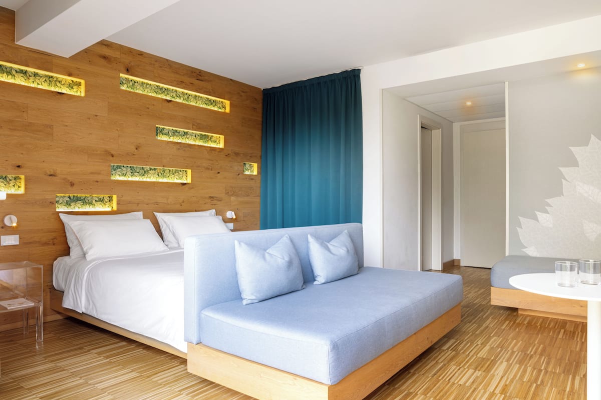 A modern hotel room with a wooden accent wall, a large bed with white bedding, a blue sofa, and a small table with glasses.