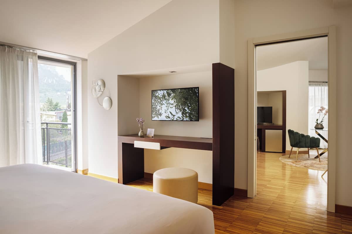 A modern hotel room with a large bed, a desk with a flat-screen TV, a small stool, and a view of the outdoors.