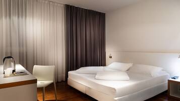 A hotel room with a bed, a chair, a desk with a coffee maker, and a nightstand with a lamp.