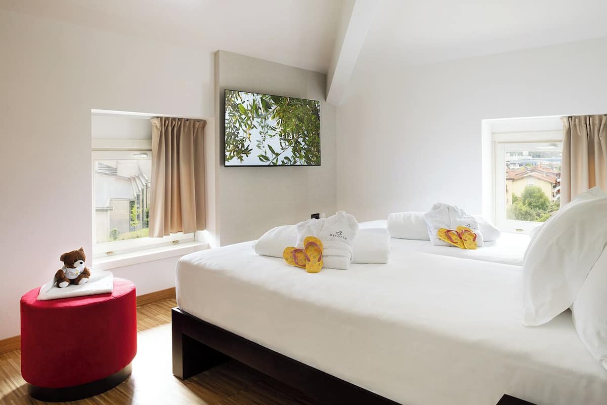 A modern hotel room with a large bed, a red ottoman, and a view of the city.