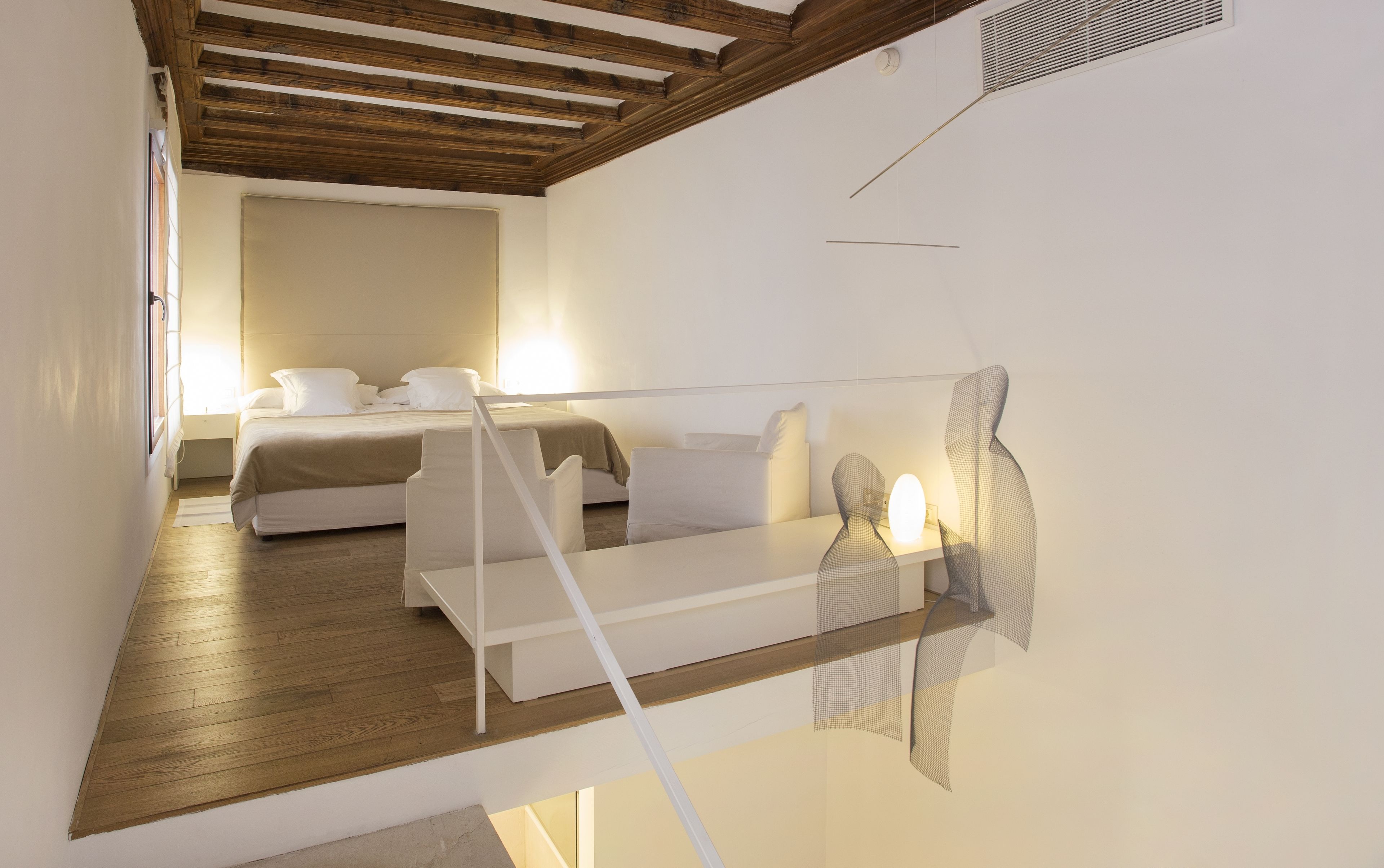 Una habitación de hotel moderna con techo de madera, cama con ropa blanca, sofá y una obra de arte original en la pared.