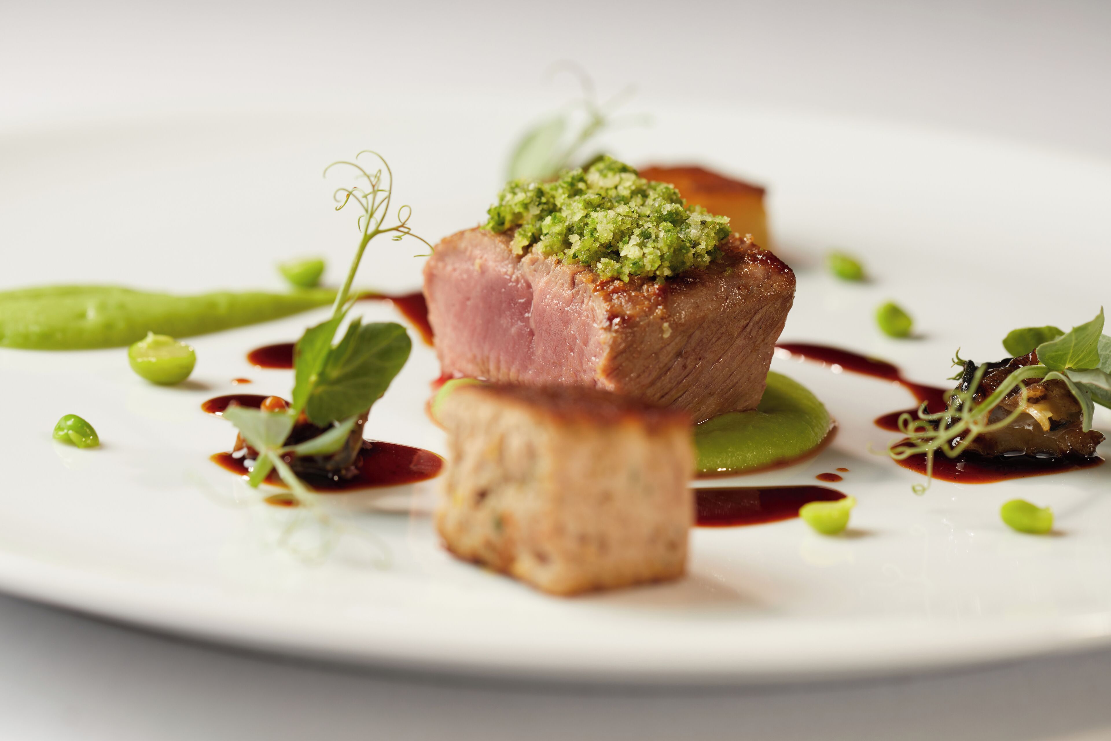 Un plato con un trozo de carne cubierto de guarnición verde, rodeado de verduras verdes y salsa.
