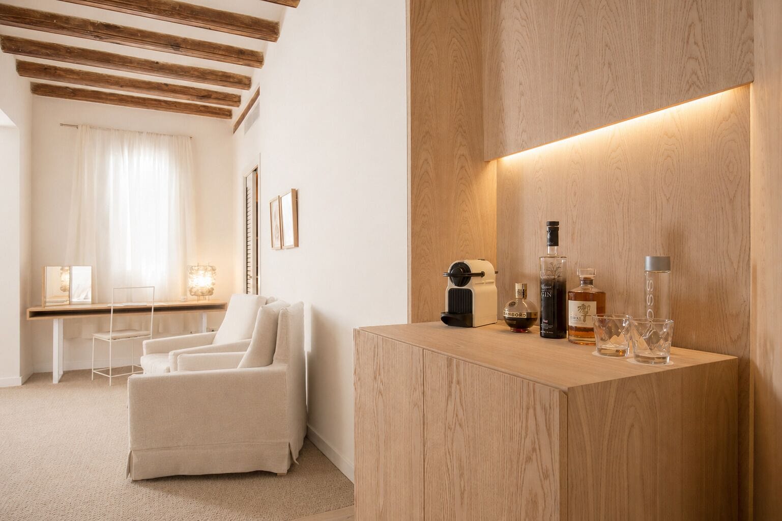 Habitación de hotel moderna con mostrador de recepción de madera, un sillón blanco y un escritorio con lámpara y marcos de fotos.