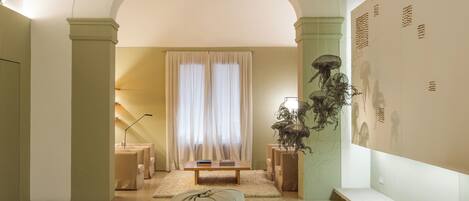A spacious living room with a high ceiling, wooden beams, a large window with curtains, and a wall-mounted artwork.