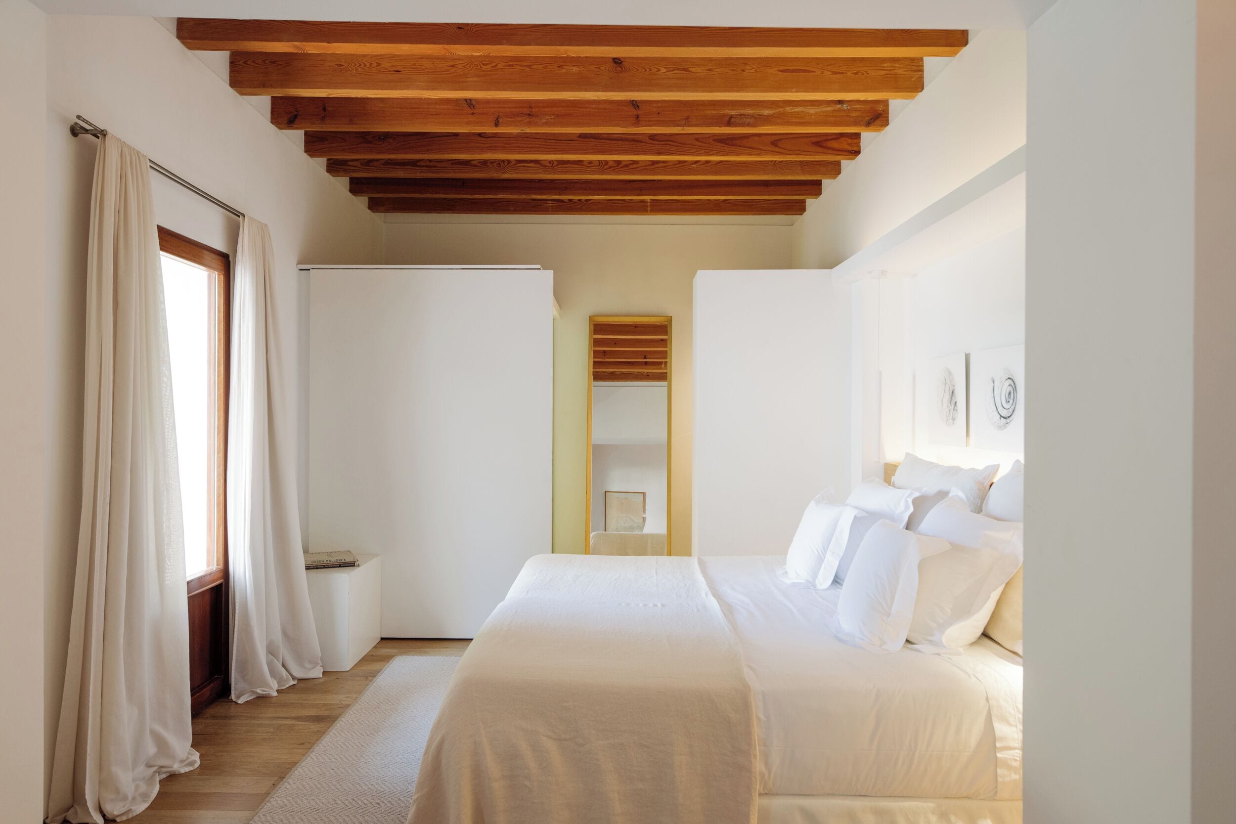 A bedroom with a wooden ceiling, a bed with white bedding, a window with curtains, and a white wardrobe.