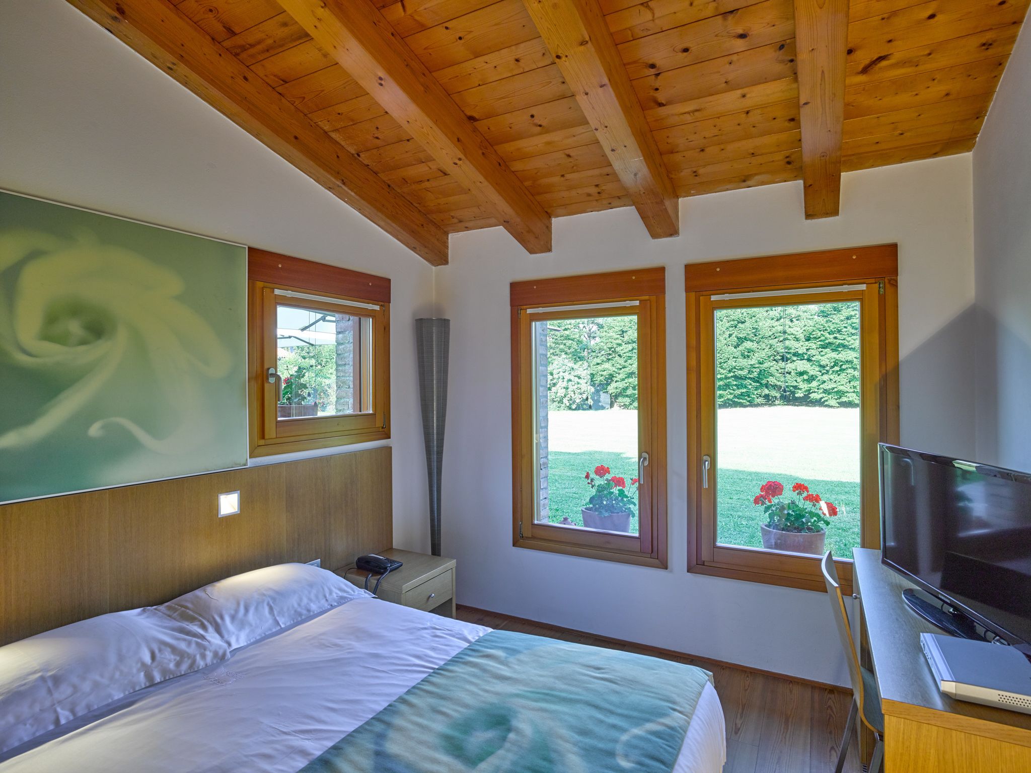 A bedroom with a bed, a desk with a TV, two windows with views of greenery, and a wooden ceiling.