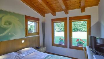 A bedroom with a bed, a desk with a TV, two windows with views of greenery, and a wooden ceiling.