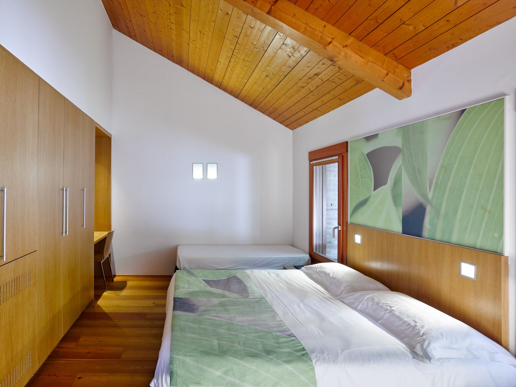 A bedroom with a wooden ceiling, a bed with a green and white bedspread, a wooden wardrobe, and a window with a plant view.