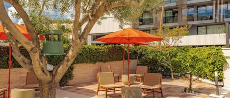 An outdoor seating area with wicker chairs, a large orange umbrella, and a tree.