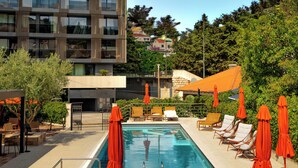 A rooftop pool area with lounge chairs and umbrellas, surrounded by greenery and a modern building. - Hotel Aquarius Adults Only (Dubrovnik)