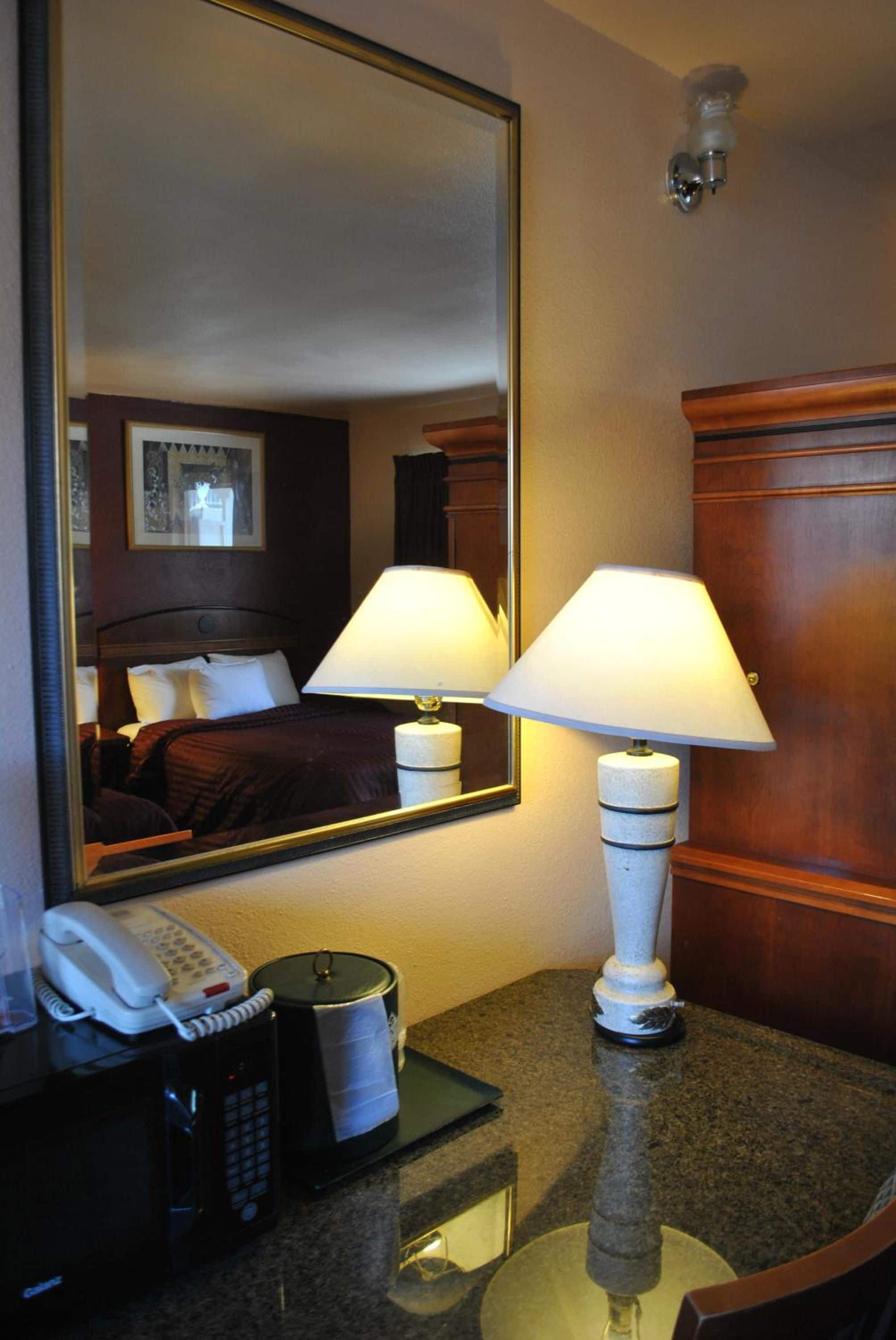 A hotel room with a bed, two bedside lamps, a telephone, a kettle, and a mirror reflecting the room.