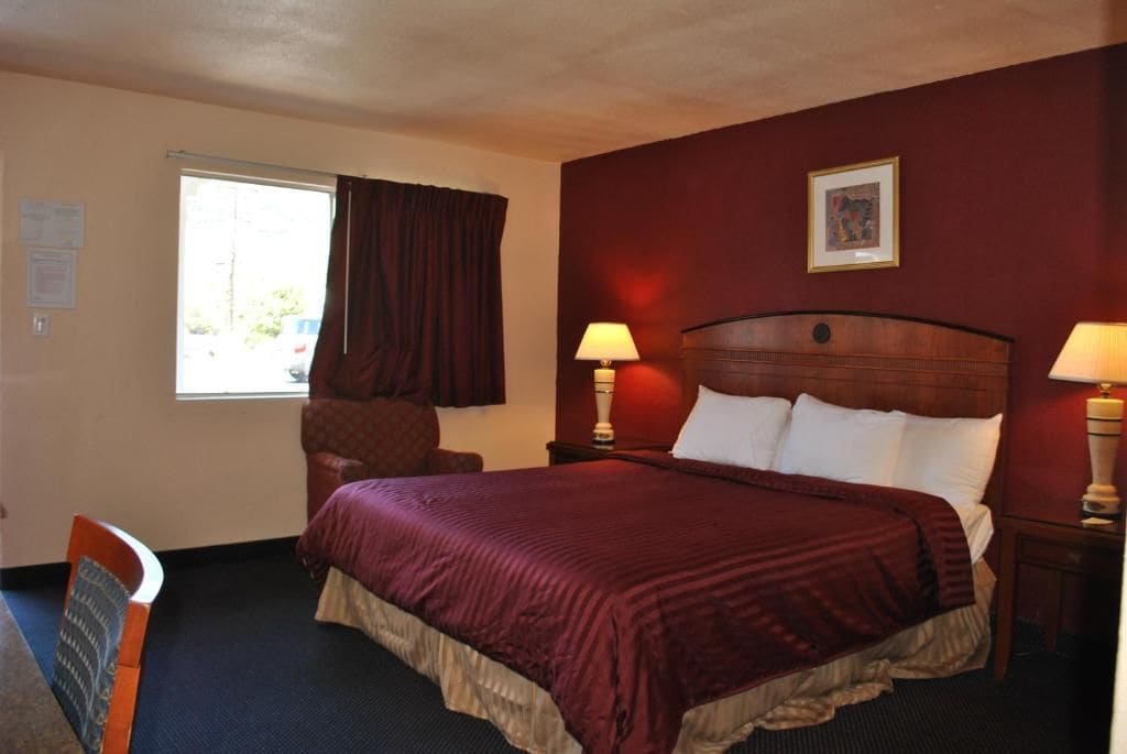 A hotel room with a large bed, two bedside lamps, a chair, a window with curtains, and a framed picture on the wall.