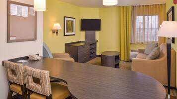 Habitación de hotel con mesa de comedor, sillas, sofá, televisor, lámpara y un tablón de anuncios.