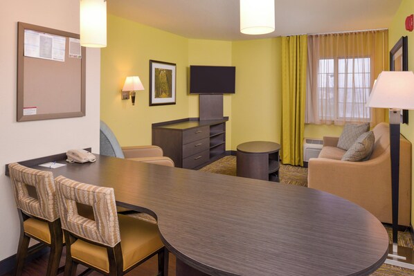 A hotel room with a dining table, chairs, a sofa, a TV, a lamp, and a bulletin board.