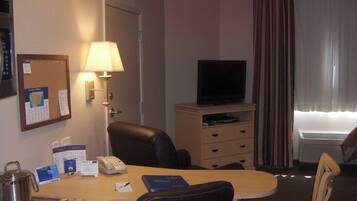 A hotel room with a desk, a chair, a television on a dresser, a lamp, and a window with curtains.