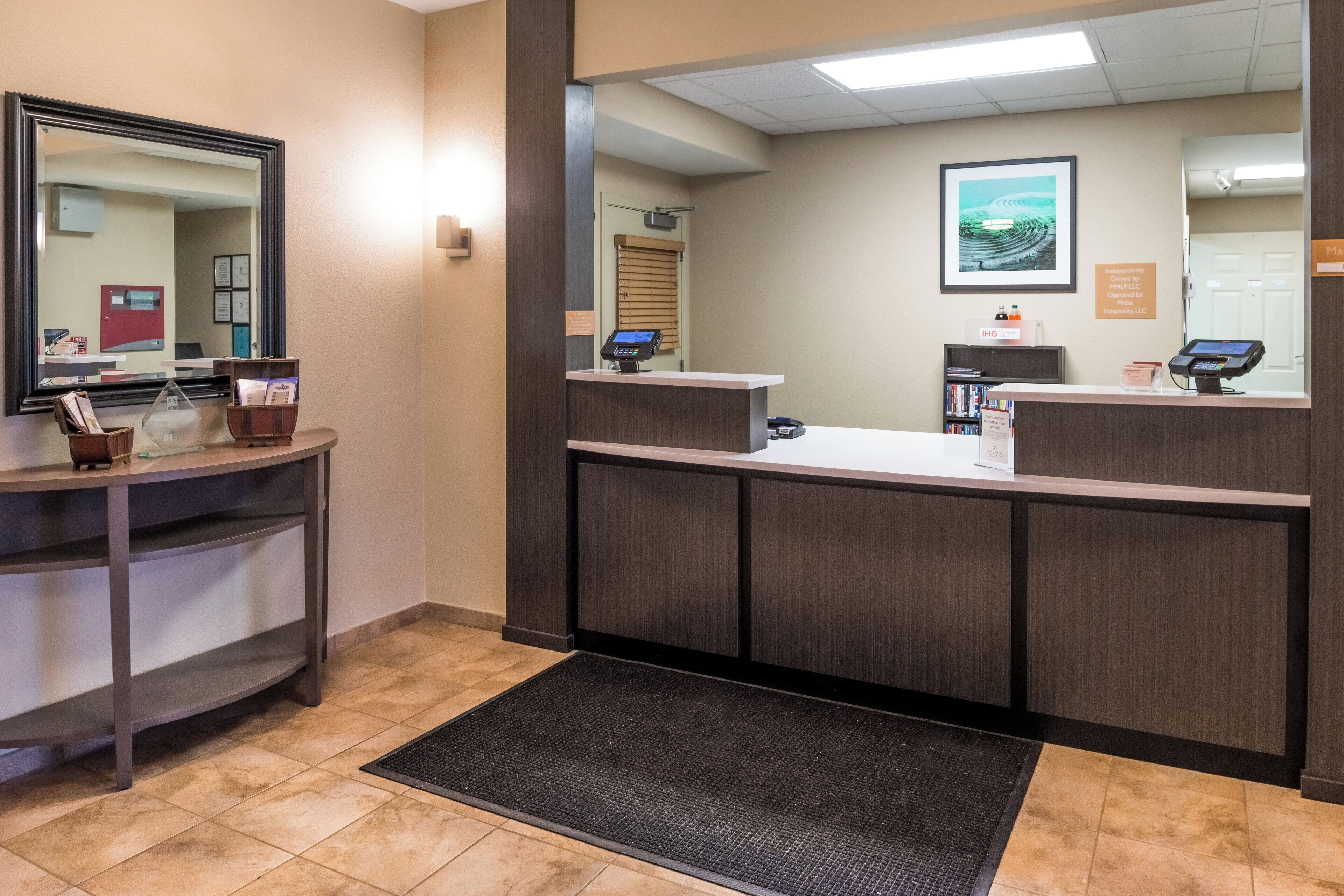 A reception area with a reception desk, a large mirror, a digital kiosk, and a small waiting area with a bench and magazines.