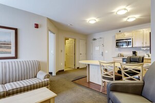 Un espacio habitable compacto con sofá, una mesa de comedor con sillas, una kitchenette y una puerta que da a otra habitación.