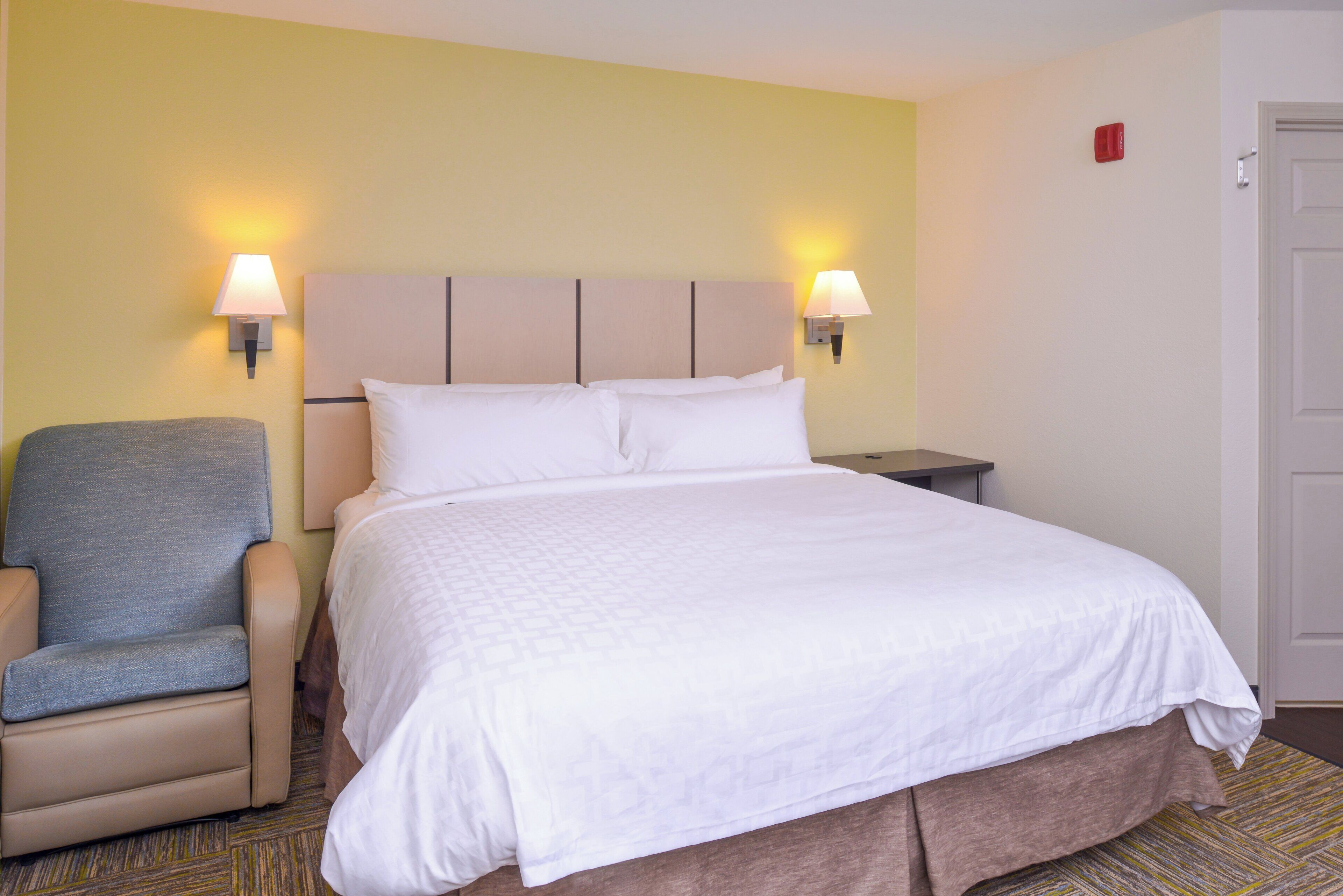 Une chambre d’hôtel comprenant un lit, une chaise, une table de chevet et deux lampes fixées au mur.