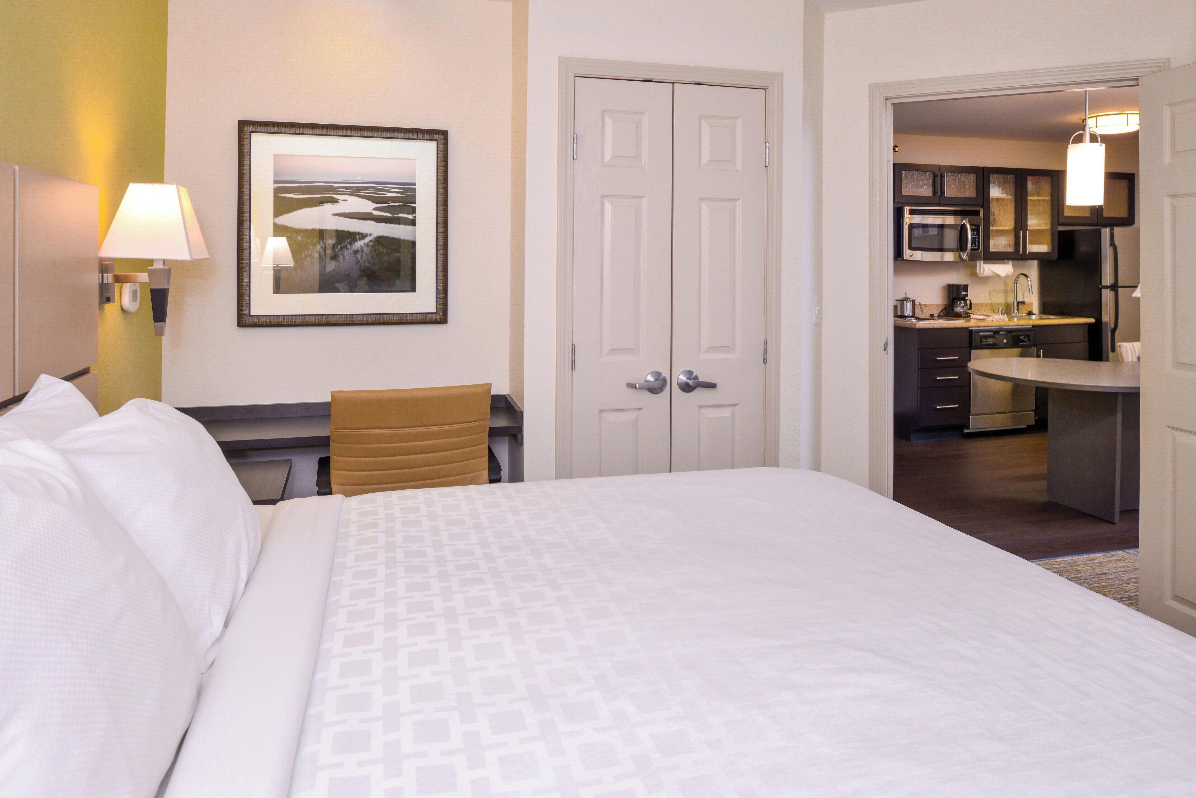 A hotel room with a bed, a desk, a chair, a framed picture, a door, and a kitchenette.