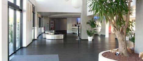Um lobby de hotel moderno com uma recepção, uma grande árvore em vaso e uma placa com o nome do hotel.