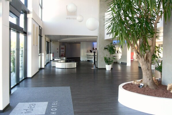 A modern hotel lobby with a reception desk, a large potted tree, and a sign indicating the hotel's name.
