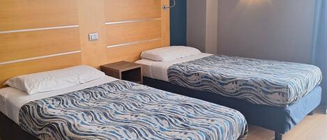 A hotel room with two beds, wooden headboards, a nightstand, a wall-mounted light, and a wooden floor.