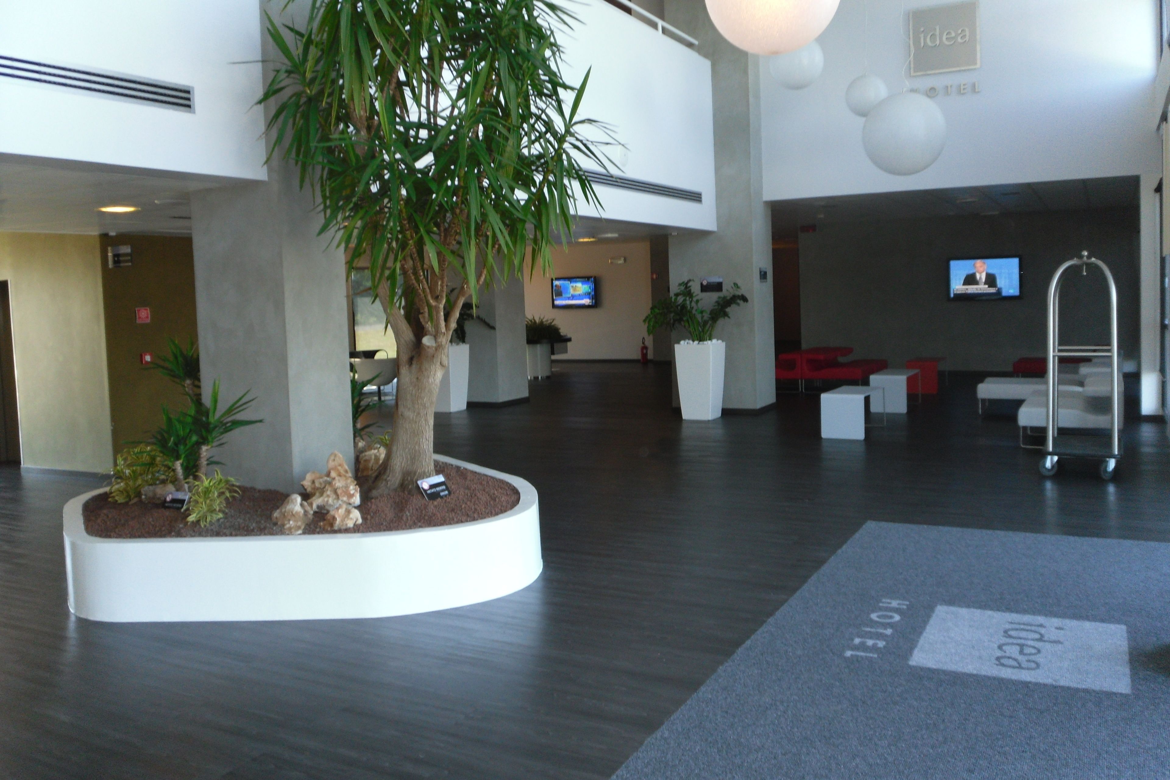Een moderne hotel lobby met een centraal geplaatste plantenbak, zithoeken en een televisiescherm.