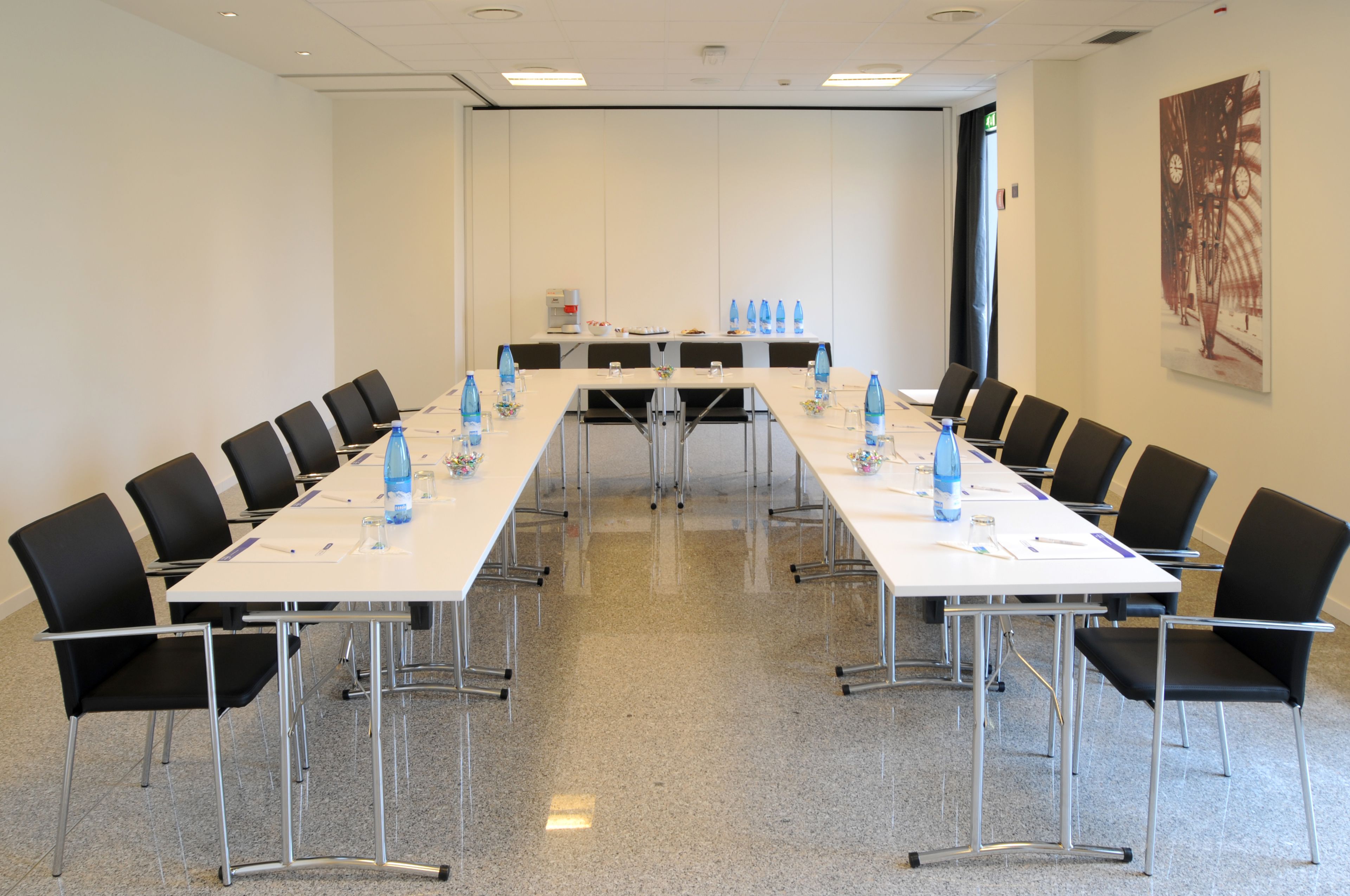 Ein Konferenzraum mit langem Tisch, schwarzen Stühlen, Wasserflaschen und einer Kaffeemaschine.