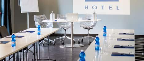 A conference room with a long table, chairs, a laptop, and water bottles. A projector screen displays "idea HOTEL".