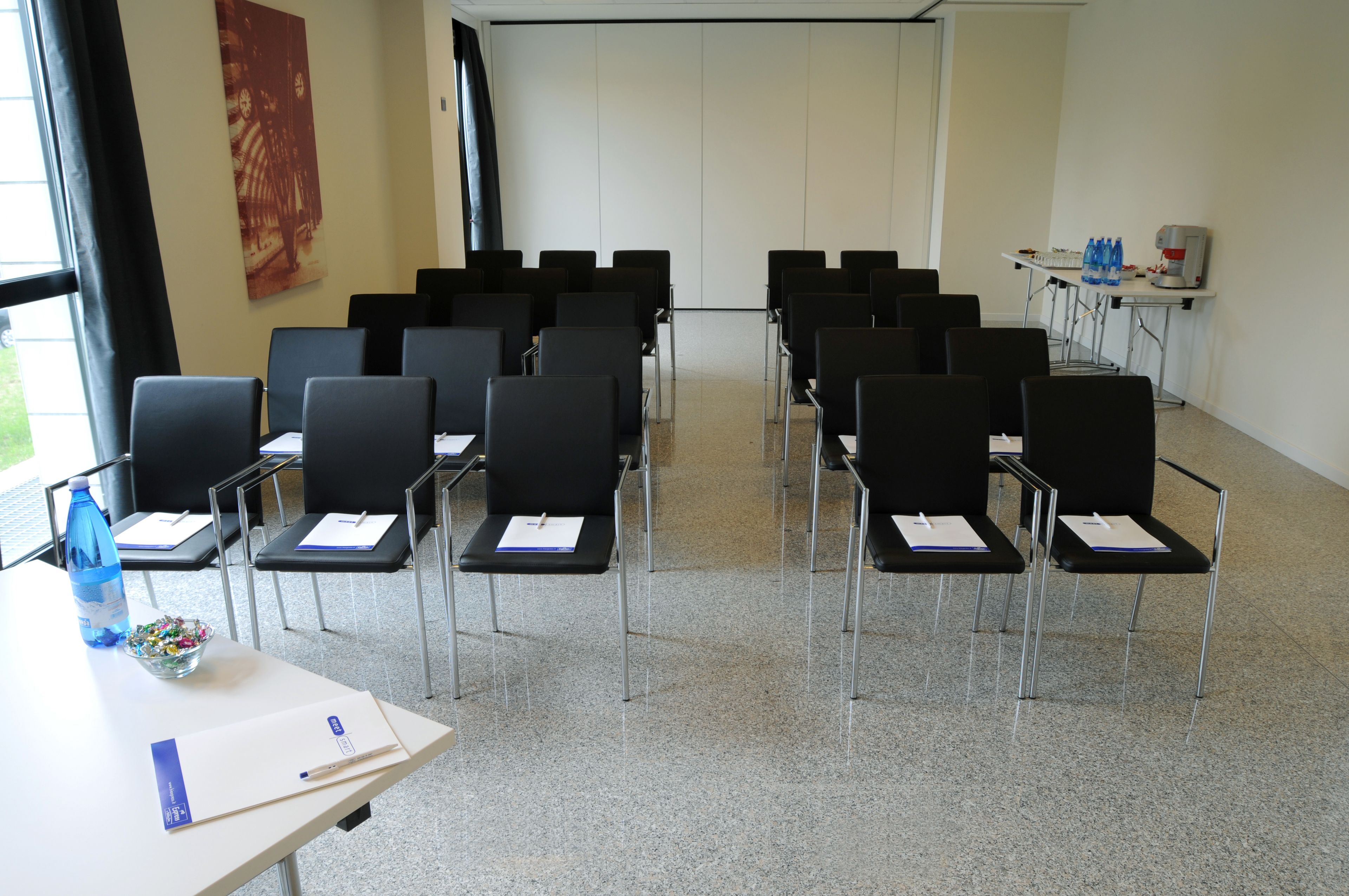 A conference room with rows of black chairs, a white table with a water bottle and papers, and a projector screen.