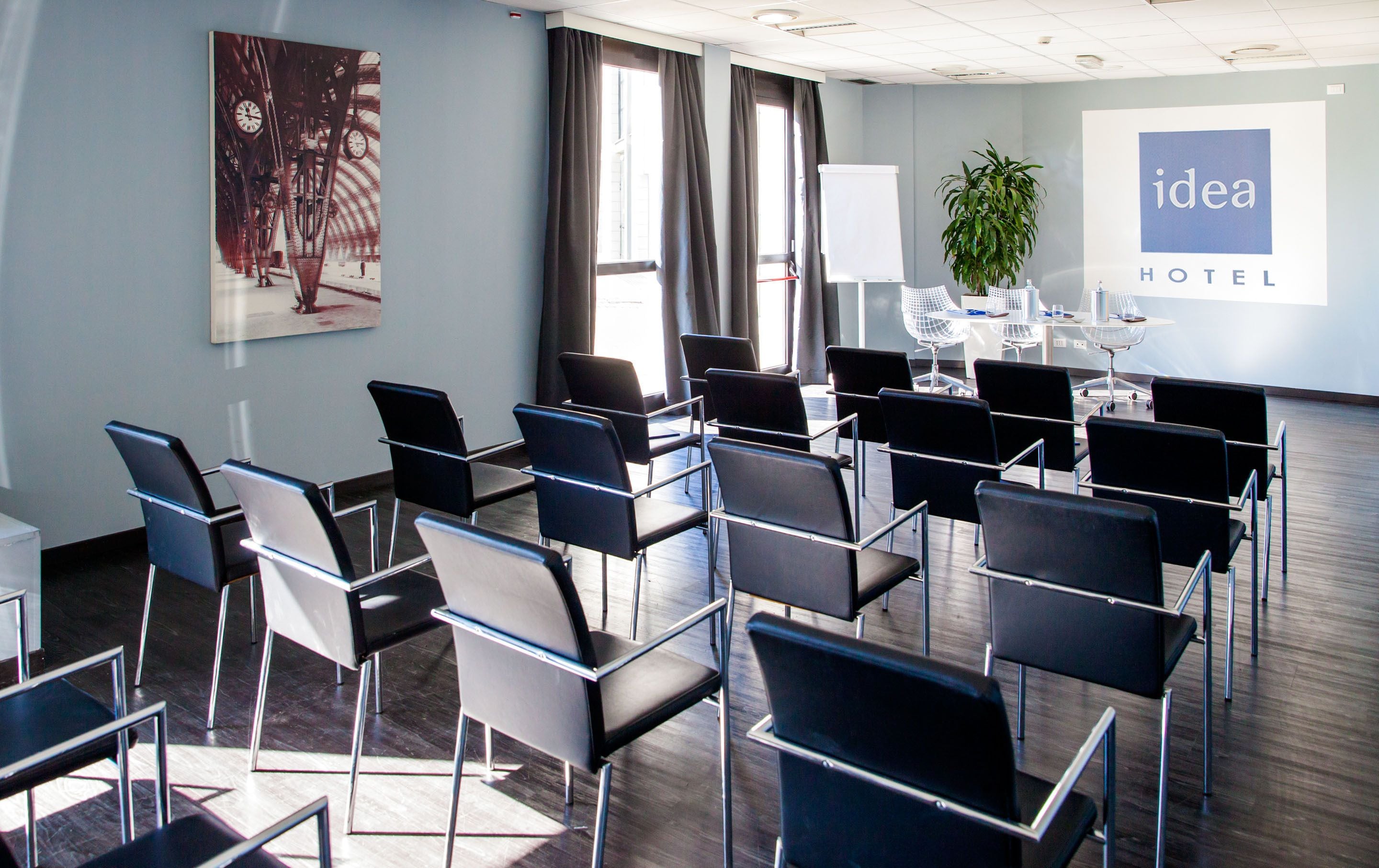 Un salón de conferencias con filas de sillas negras, un cuadro grande, una pantalla de proyector y una planta en maceta.