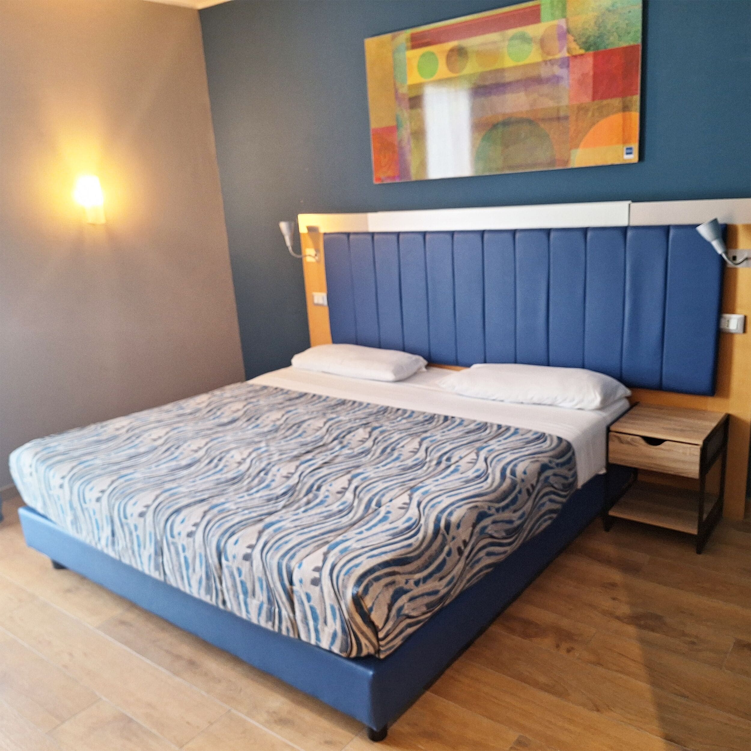 Un dormitorio con cama, mesita de noche, lámpara de pared y una obra de arte colorida sobre la cama.
