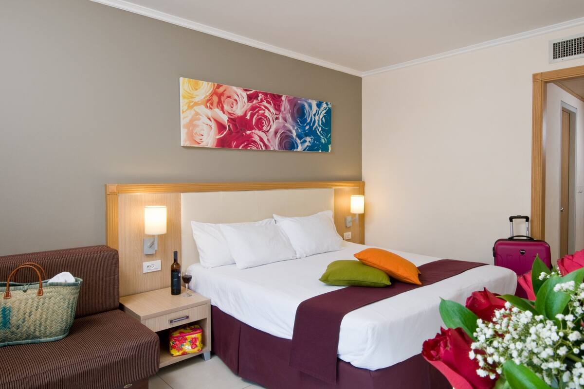 Een hotelkamer met een bed, nachtkastje, lamp, koffer en een schilderij aan de muur.