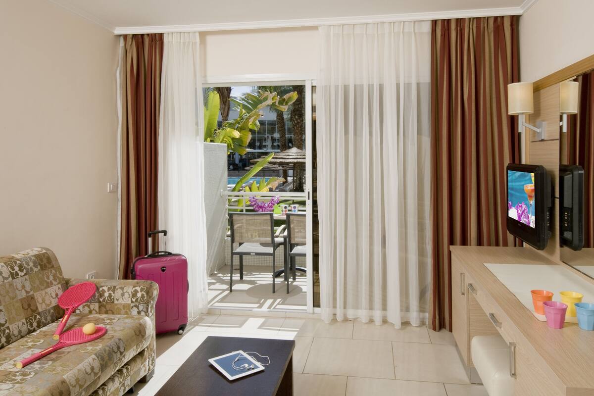 Hotelkamer met een bank, een klein tafeltje met een tablet en koptelefoon, een roze koffer, een televisie en een balkon met uitzicht op een zwembad en palmbomen.