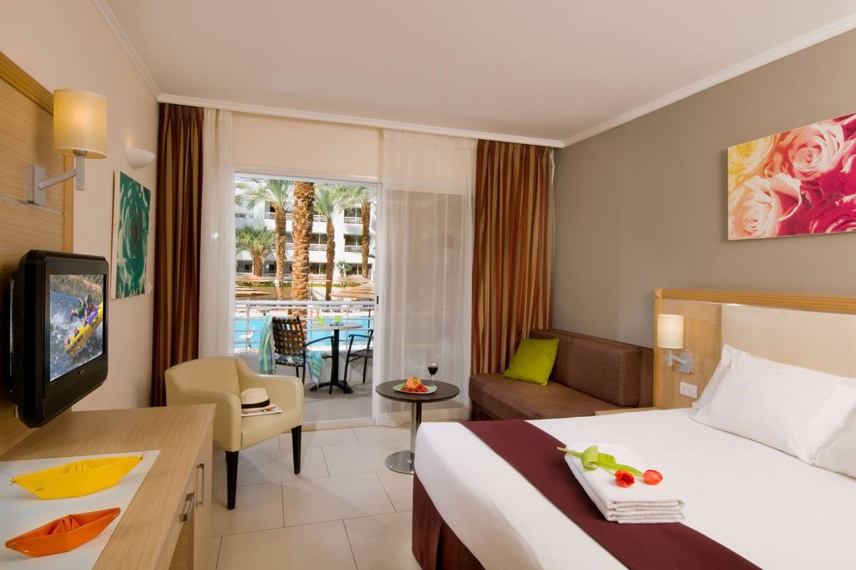 Hotelkamer met een bed, een televisie, een eettafel met stoelen, een sofa, een klein balkon met uitzicht op het zwembad, en een schilderij aan de muur.