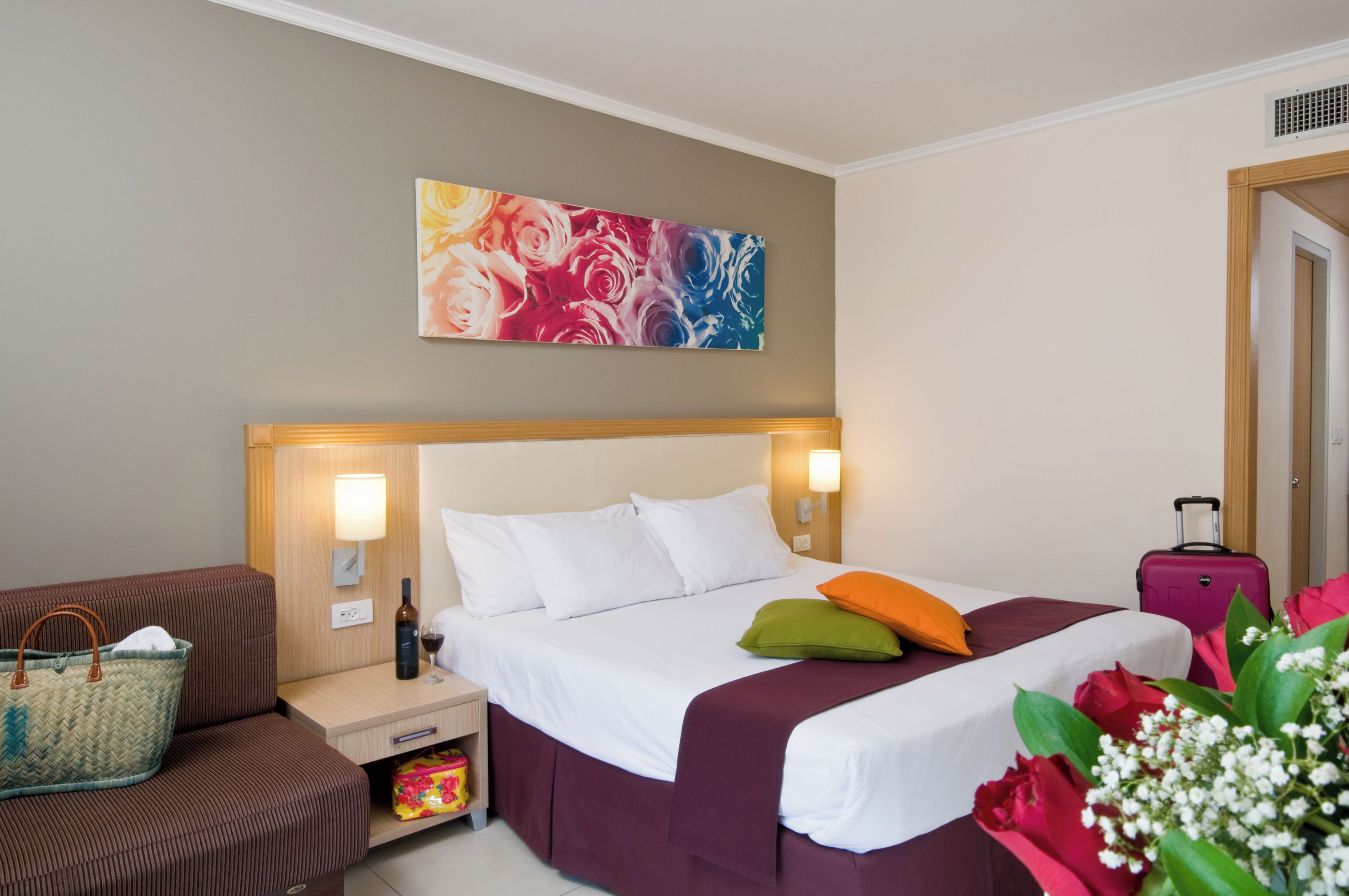 A hotel room with a bed, bedside table, lamp, and a colorful abstract painting on the wall.