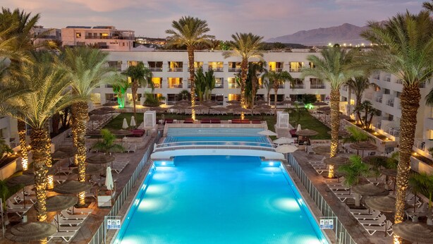 A swimming pool with lounge chairs and umbrellas, surrounded by palm trees and a hotel building. - Leonardo Royal Resort Hotel Eilat (Eilat)