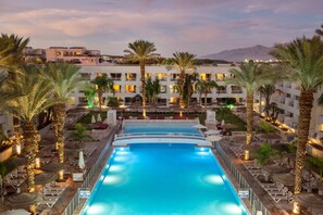 Una piscina con sillones de descanso y sombrillas, rodeada de palmeras y un edificio de hotel.