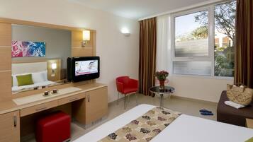 A hotel room with a bed, a desk, a TV, a red chair, a small table with a vase, and a window with curtains.