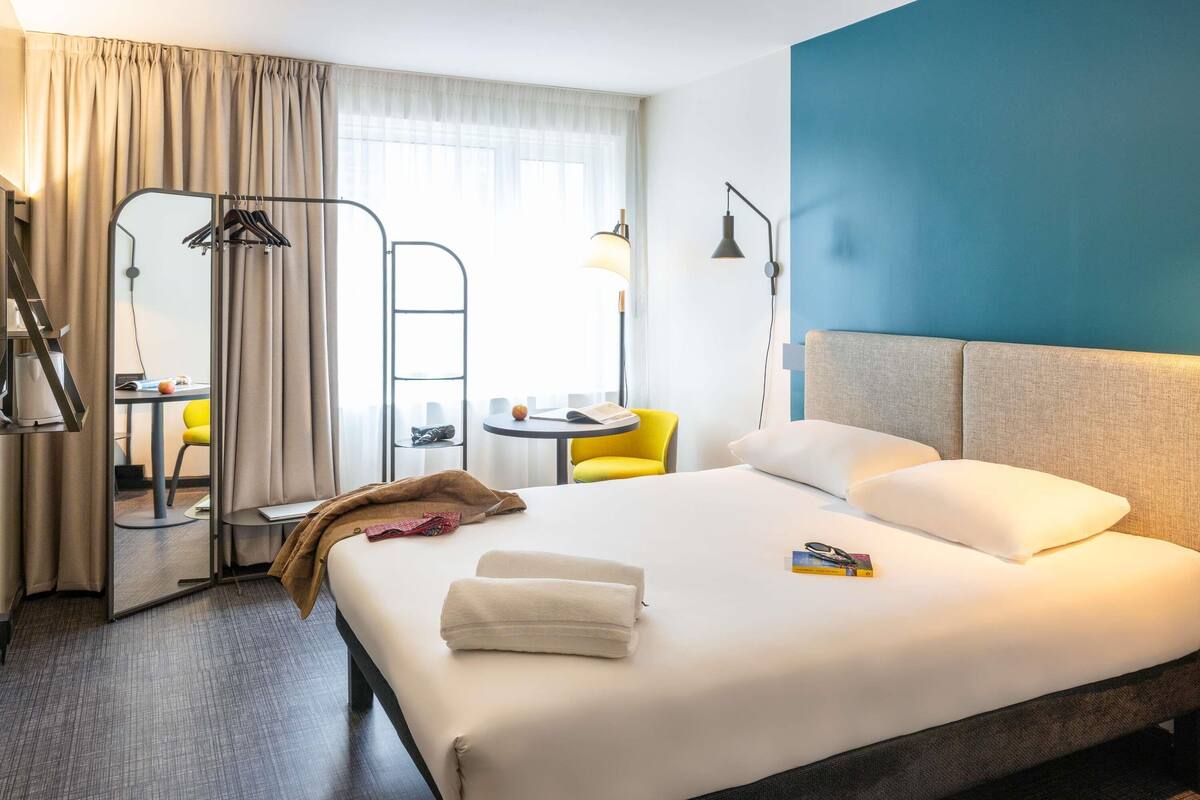 A hotel room with a bed, a desk, a chair, a lamp, and a view of the city.