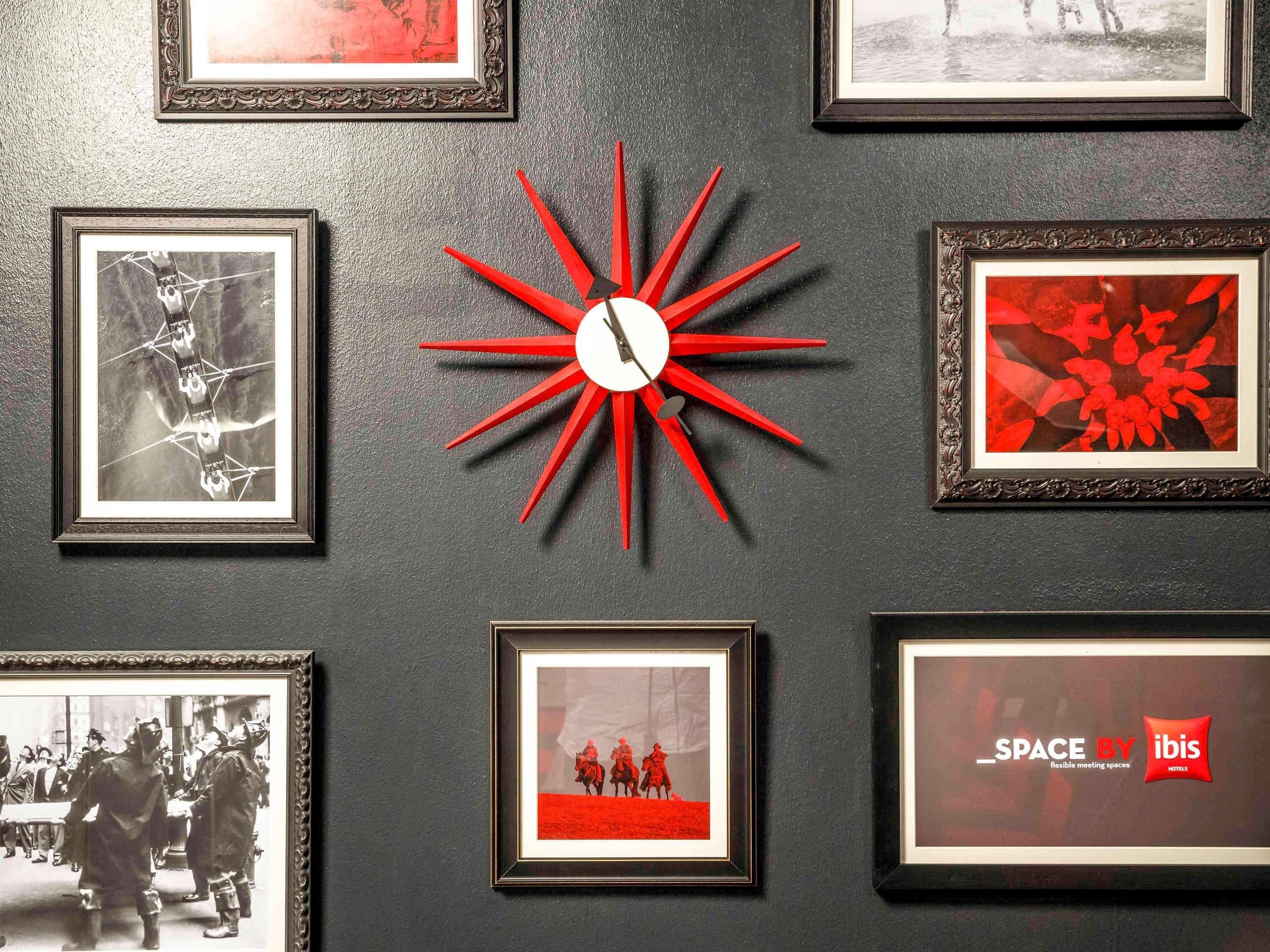 A wall with multiple framed pictures, a central red clock, and a sign for 'SPACE BY ibis Hotels'.