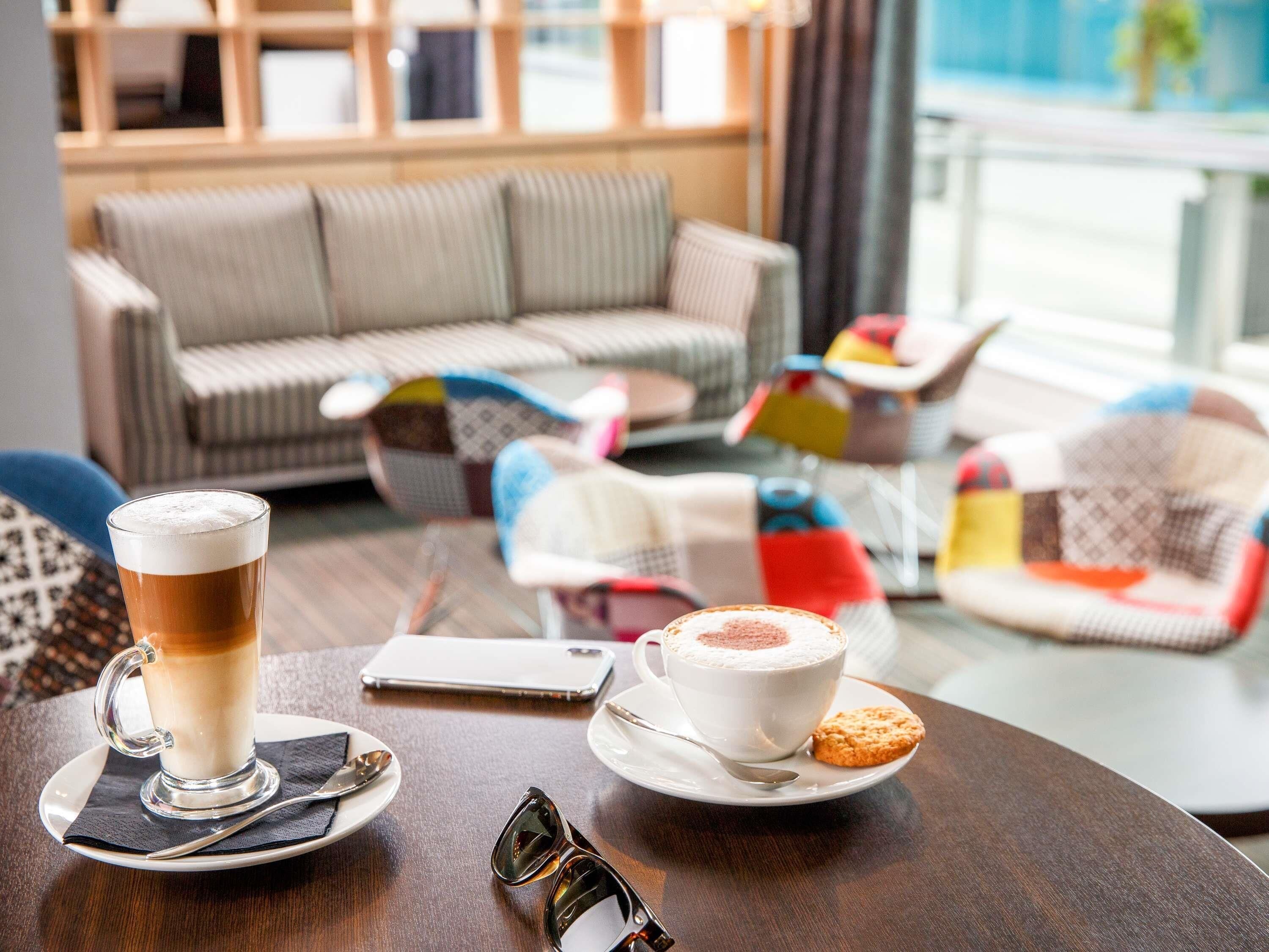 Un rincón acogedor con una mesa dispuesta para dos, que incluye una taza de café, una de té, un par de gafas de sol y un teléfono inteligente.