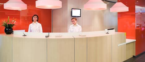 A reception desk with two staff members, a modern interior, and a flat-screen TV on the wall.