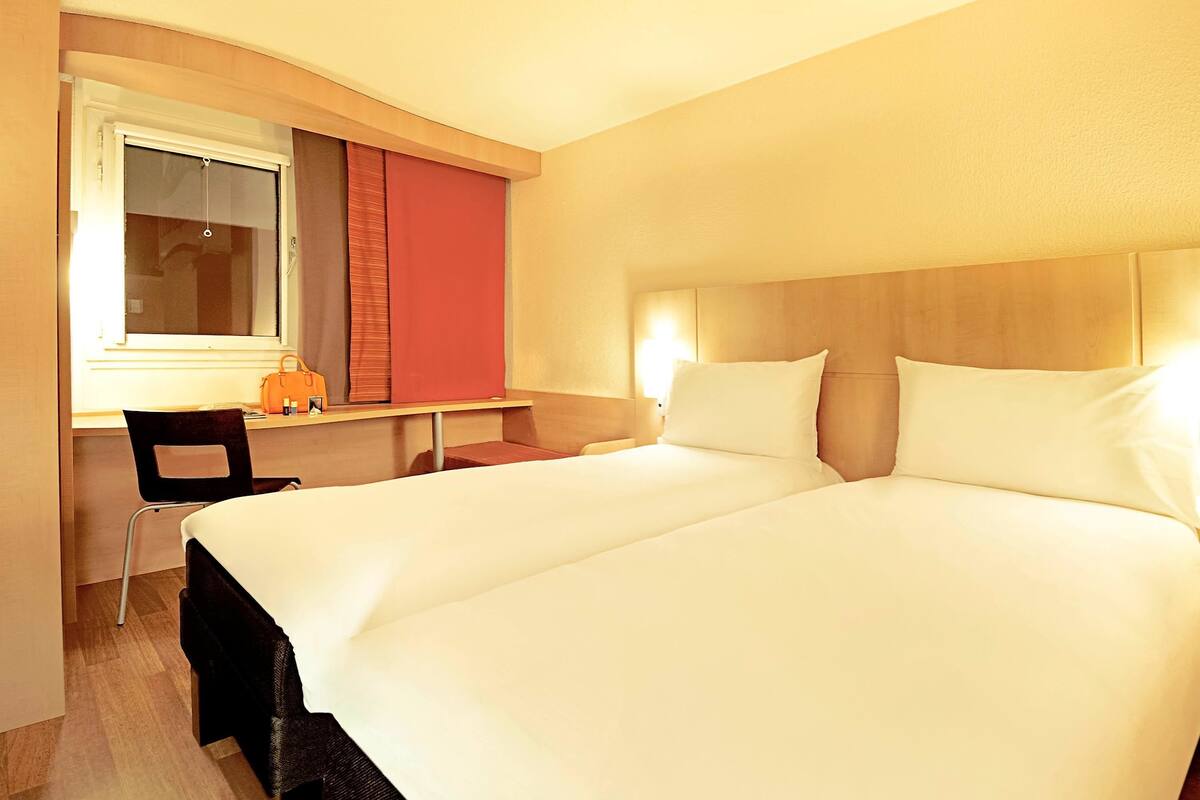 A hotel room with a double bed, a desk with a chair, and a window with curtains.